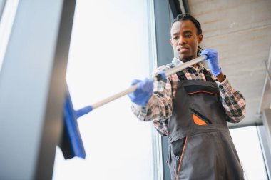 Mavi eldiven giyen ve cam sileceği kullanan Afro-Amerikan temizlik işçisi ofis binasının camlarını temizliyor.
