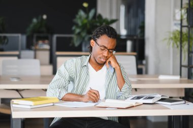 Genç Afrikalı Amerikalı öğrenci, kitaplar ve öğrenim kaynaklarıyla çevrili, parlak, modern bir üniversite kütüphanesinde özenle yazıyor ve okuyor.