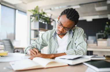 Genç Afrikalı Amerikalı erkek öğrenci kütüphanede çalışırken ders kitaplarından notlar alıyor.