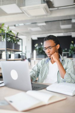 Genç Afrikalı Amerikalı öğrenci araştırma için dizüstü bilgisayar kullanırken konsantre oluyor ve parlak üniversite kütüphanesinde internetten öğreniyor.