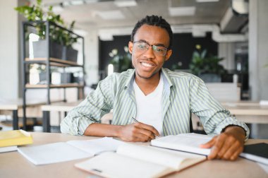 Genç Afrikalı Amerikalı öğrenci üniversite kütüphanesinde okuyor, ders kitaplarından notlar alıyor ve sınavlara hazırlanıyor.