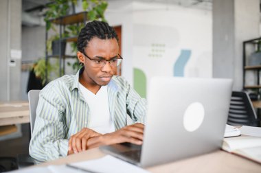 Genç adam dizüstü bilgisayarına odaklanmış, modern bir üniversite kütüphanesinde proje üzerinde çalışıyor.