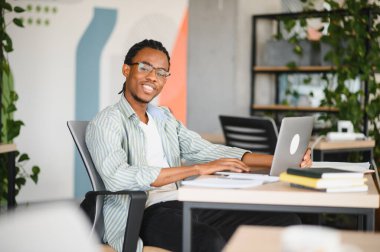 Bilgisayarda çalışan, sınavlara hazırlanan, üniversite kütüphanesinde ödev yapan, etrafı kitaplarla çevrili mutlu Afrikalı Amerikalı öğrenci.