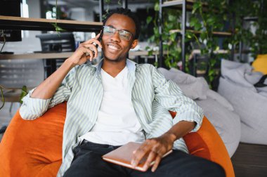 Mutlu Afrikalı Amerikalı öğrenci, modern bir üniversite kütüphanesinde armut koltukta dinlenirken bir telefon görüşmesi yapıyor ve elinde bir defter tutuyor.