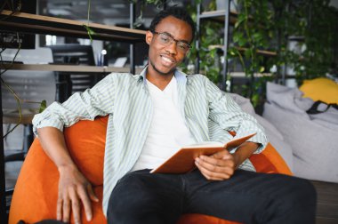 Genç siyahi bir adam yüksek öğrenim ve akademik başarıyı temsil eden rahat bir ortamda kitap okurken derslerinden zevk alıyor.