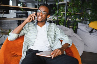 Gülümseyen Afrikalı Amerikalı öğrenci üniversitede okurken telefonda konuşuyor. Bir kitap tutmak, rahat, bitki dolu bir çevreyle çevrili.