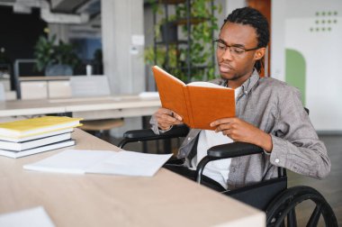 Özürlü bir genç adam özenle çalışıyor ve modern, parlak bir ofiste kitap okuyor. İçi doğal ışık ve modern mobilya dolu.