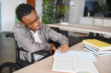 Tekerlekli sandalye kullanan, parlak, modern ofis ya da kütüphanede okuyan ve not alan engelli genç siyahi bir adam.