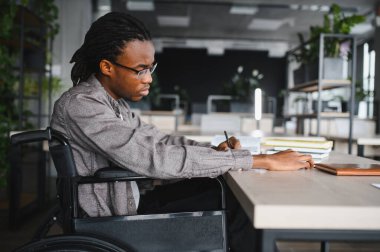 Modern üniversite kütüphanesinde masa başında çalışırken tekerlekli sandalyeyle yazı yazan genç adam, eğitim ve akademik başarıya odaklandı