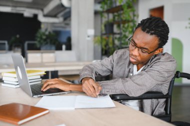 Tekerlekli sandalye kullanan genç siyah adam özenle çağdaş bir ofiste çalışıyor, notlar alıyor ve bir dizüstü bilgisayar kullanıyor, kendini işine adadığını ve odaklandığını gösteriyor.