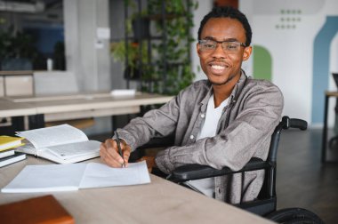 Tekerlekli sandalyedeki gülümseyen Afrikalı Amerikalı öğrenci modern bir sınıftaki bir masaya notlar yazıyor, eğitimi ve ulaşılabilirliği vurguluyor.