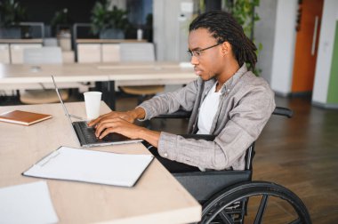 Modern ofiste dizüstü bilgisayarla çalışan engelli genç profesyonel bir adam, iş yerindeki dahil olma ve erişilebilirliği gösteriyor