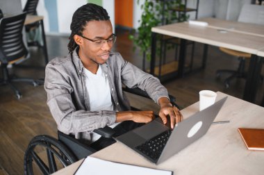 Tekerlekli sandalyedeki genç adam dizüstü bilgisayarla çalışmaya odaklanmış engelliler için erişilebilirliği ve eğitimi sembolize ediyor.