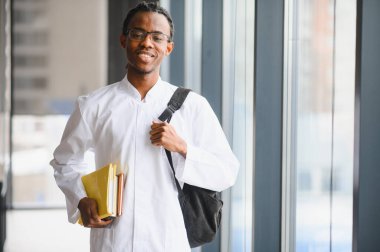 Beyaz önlüklü, kitap ve sırt çantası taşıyan, üniversite koridorunda gülümseyen genç siyah erkek öğrenci.