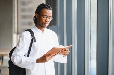 Üniversite koridorundaki büyük bir pencerenin yanında kitap okuyan odaklanmış tıp öğrencisi, sınavlara hazırlanıyor ve bilgiyi genişletiyor.