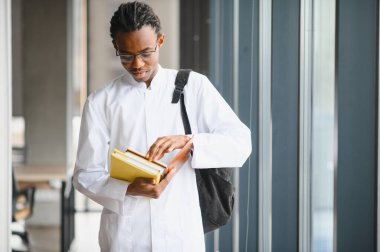 Laboratuvar önlüğü ve sırt çantası giymiş, parlak bir üniversite koridorunda kitap okuyan odaklanmış Afrikalı Amerikalı öğrenci.