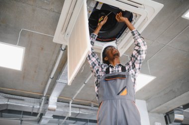 Afro-Amerikan hvac teknisyeni tulum ve başlık takıyor. Havalandırma sistemini onarmak için tornavida kullanıyor.