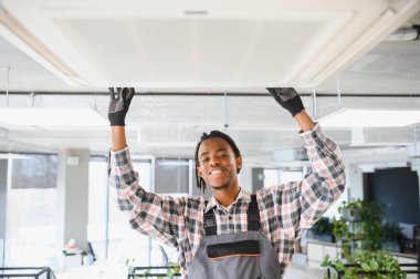 Afrika kökenli Amerikalı teknisyen bir ofis havalandırma sistemini ustalıkla onarıyor ve verimli havalandırma sağlıyor. Doğal ışık alan kapalı bir ortam.
