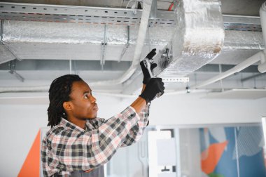 Genç Afro-Amerikan işçiler klima borularına kilit vuruyor, modern ofis ortamında bakım yapıyorlar.