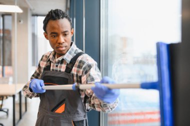 Genç Afro-Amerikan adam mavi eldiven giyiyor ve gri tulumla camları siliyor.