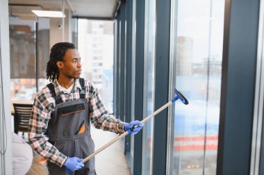 Genç adam modern bir ofis binasının camlarını temizlemek için teleskopik cam sileceği kullanıyor. Temiz ve hijyenik bir çalışma ortamı sağlıyor.