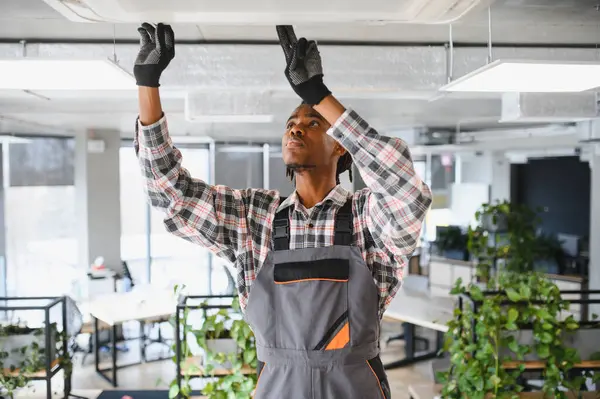 Genç Afro-Amerikan bakım işçisi modern ofiste eldiven ve tulum giyerek havalandırma sistemini tamir ediyor.