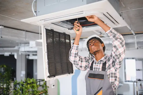 Afro-amerikan hvac teknisyeni sıkı bir şapka ve tulum giyerek bir ofis binasının tavanına monte edilmiş klima ünitesini tamir etmek için tornavida kullanıyor.