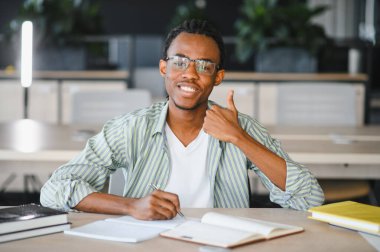 Mutlu Afro-Amerikan üniversite öğrencisi modern kütüphanede masa başında otururken notlar alıyor ve işaret ediyor.