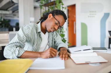 Genç erkek öğrenci, kitaplarla çevrili üniversite kütüphanesinde notlar yazıyor ve özenle ders çalışıyor ve akademik başarıya odaklanıyor.