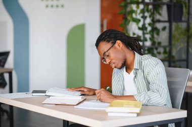 Genç adam kütüphanede ders kitapları okurken notlar yazıyor, sınavlara ve mezuniyete hazırlanıyor.