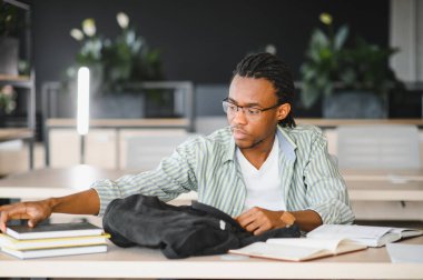 Odaklanmış Afro-Amerikan üniversite öğrencisi kütüphanede okurken sırt çantasından kitap ve defter çıkarıyor.