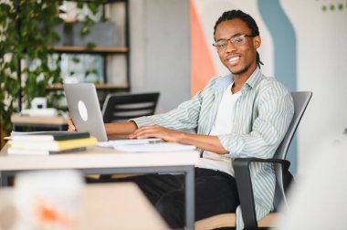 Genç Afro-Amerikan erkek öğrenci laptopuyla parlak bir üniversite kütüphanesinde kitap ve evrak işleriyle çalışıyor.
