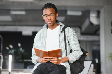 Genç Afrikalı Amerikalı erkek öğrenci, kitap okuyor, gözlüklü ve sırt çantalı, modern bir üniversite kütüphanesinde, sınavlara hazırlanıyor.