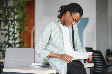 Odaklanmış Afro-Amerikan üniversite öğrencisi ders kitabı okuyor, kütüphanede sınavlara hazırlanıyor.