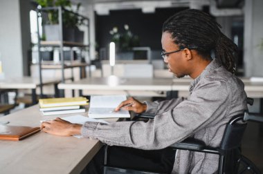 Tekerlekli sandalye kullanan engelli genç adam, birlikte çalışma alanında öğreniyor, not defterine not alıyor.