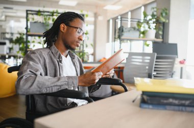 Tekerlekli sandalye kullanan genç bir adam, parlak ve modern bir ofis ortamında çalışırken kitap okuyor.