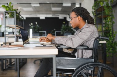 Masa başında dizüstü bilgisayar kullanan engelli bir adam, modern, bitki dolu bir çalışma alanında çalışmaya odaklanmış. Eğitimi ve erişilebilirliği vurguluyor