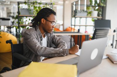 Özürlü genç adam dizüstü bilgisayar kullanıyor ve modern ofiste notlar alıyor, kapsamlı eğitim ve işyeri çeşitliliğini kucaklıyor