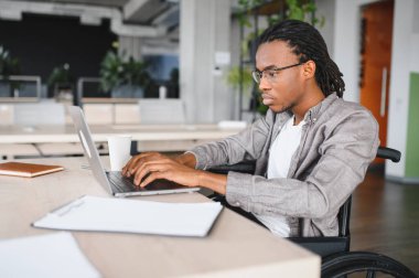 Tekerlekli sandalyedeki Afrikalı Amerikalı öğrenci modern ofis ortamında dizüstü bilgisayar üzerinde çalışıyor, eğitimde çeşitlilik ve erişilebilirliği vurguluyor