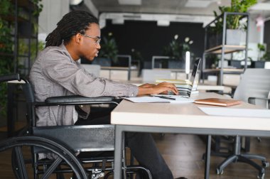 Özürlü genç bir adam, modern ofiste dizüstü bilgisayarla çalışıyor. İşyerine dahil edilme ve erişilebilirlik gösteriyor.