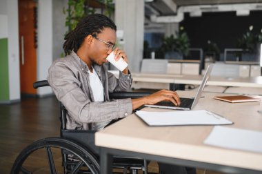 Modern bir ofiste dizüstü bilgisayarla çalışırken kahveden zevk alan engelli genç bir adam azmini ve kariyerine odaklanmasını gösteriyor.