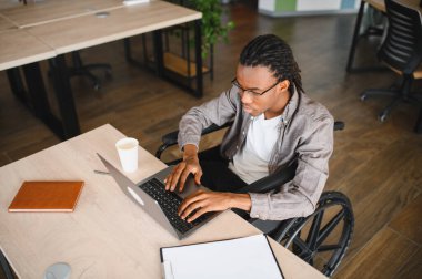 Tekerlekli sandalye kullanan, engelli genç bir Afrikalı Amerikalı, modern bir ofiste, bilgisayarının başında, görevlerine odaklanmış bir şekilde çalışıyor.