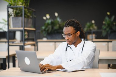 Modern ofis ortamında dizüstü bilgisayar kullanan, bitkilerle çevrili, steteskoplu Afrikalı Amerikalı erkek doktor.