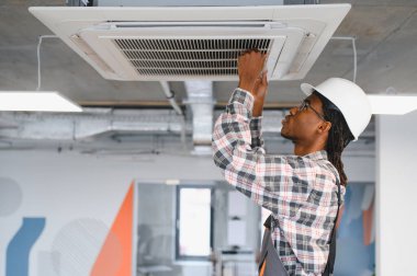 Modern bir ofis ortamında tavan monte edilmiş klimayı tamir eden sert şapkalı ve ekose gömlekli profesyonel bir teknisyen.