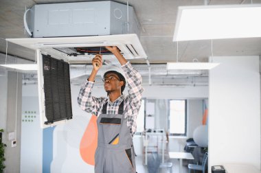 Afrikalı Amerikan hvac teknisyeni modern bir ofisin tavanındaki klimayı tamir etmek için tornavida kullanıyor.