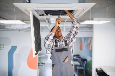 Afrikalı Amerikan hvac teknisyeni, modern ofis ortamında bakım yapan bir klima ünitesinde çalışırken tornavida kullanıyor.