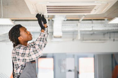 Profesyonel üniformalı ve eldivenli işçiler modern bir endüstriyel ortamda tavana monte edilmiş klima ünitesinde bakım yapıyorlar.