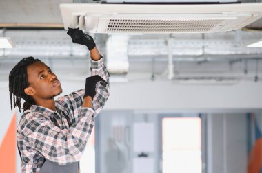 Genç Afro-Amerikan HVK teknisyeni bir binanın tavanına monte edilmiş bir havalandırmayı inceliyor ve onarıyor.