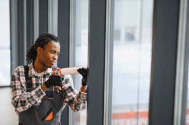 Odaklanmış Afro-Amerikan inşaat işçisi, bir iskan binasına cam yerleştirirken, bir kalıntı tabancasıyla dolgu macunu sürüyor.