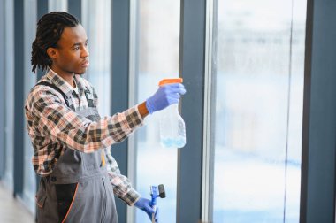 Modern bir bina ortamında bakım ve temizliğe odaklanan, sprey şişesi ve cam sileceği ile pencereleri temizleyen profesyonel bir işçi.